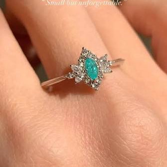 A silver-toned ring with a marquise-cut turquoise center stone and small clear stones, shown worn on a person's finger. The image has a soft-focus background and text overlays reading 'Small but unforgettable.